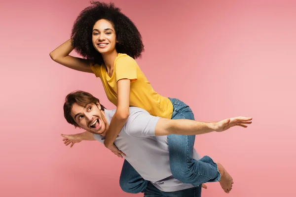 Feliz latino mujer piggybacking en asombrado novio imitando plano con extendida manos aislado en rosa - foto de stock