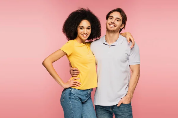 Jovem e alegre casal interracial olhando para a câmera enquanto abraça isolado no rosa — Fotografia de Stock