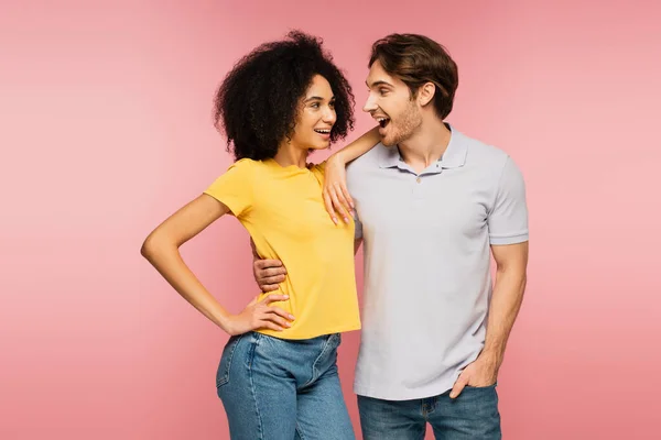 Erstaunt schaut Mann freudige lateinische Freundin an, die mit der Hand auf Hüfte isoliert auf rosa steht — Stockfoto