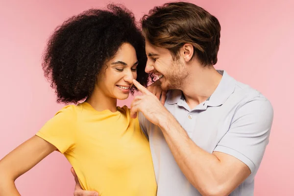 Homem alegre abraçando feliz namorada hispânica ao tocar seu nariz isolado em rosa — Fotografia de Stock