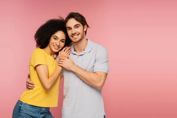 Junges und glückliches multiethnisches Paar, das sich umarmt, während es in die Kamera lächelt, isoliert auf rosa — Stockfoto