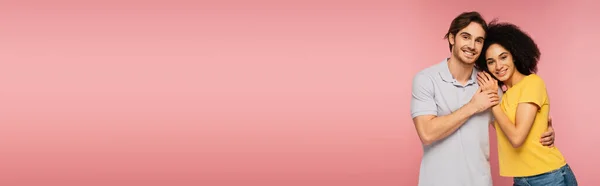 Счастливая межрасовая пара улыбается в камеру, обнимаясь изолированы на розовый, баннер — стоковое фото