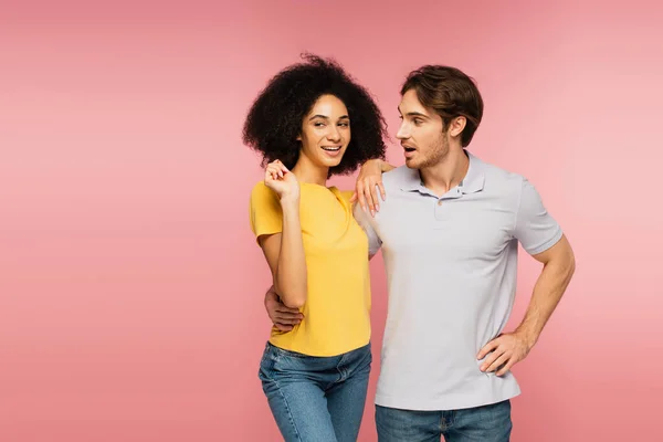 Sorprendido hombre abrazando alegre hispano mujer mientras de pie con la mano en la cadera aislado en rosa - foto de stock