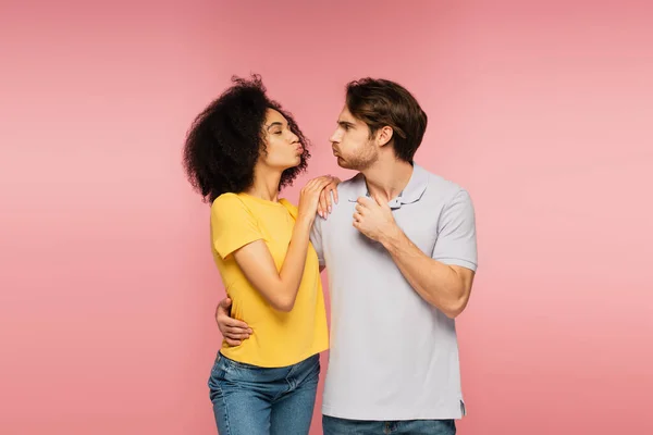 Giovane coppia multietnica abbracciare e sbuffare guance mentre si guarda l'un l'altro isolato su rosa — Foto stock