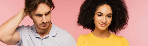 Cheerful latin woman smiling at camera near confused man touching head isolated on pink, banner — Stock Photo