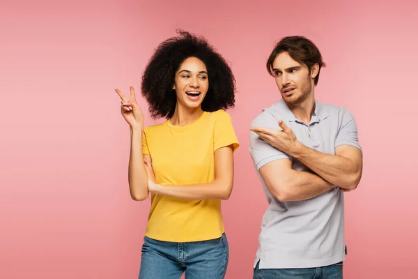 Upset man pointing at excited hispanic woman showing victory gesture isolated on pink — Stock Photo