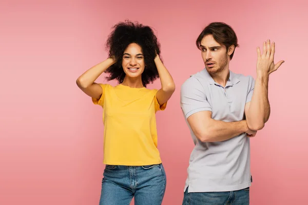 Displeased man gesturing near cheerful latin woman touching head isolated on pink — Stock Photo