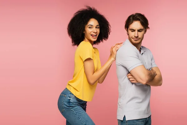 Alegre mujer latina sobresaliendo de la lengua mientras toca la espalda de un hombre molesto de pie con los brazos cruzados aislados en rosa - foto de stock