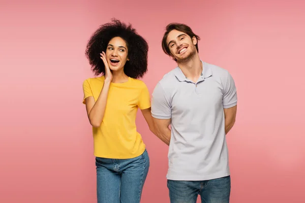 Mujer hispana asombrada tocando la cara cerca de hombre feliz ocultando algo detrás de la espalda aislado en rosa - foto de stock