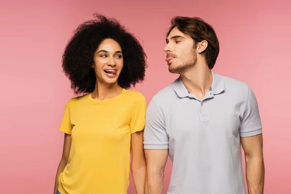 Joyful multiethnic couple looking at each other and sticking out tongues isolated on pink — Stock Photo