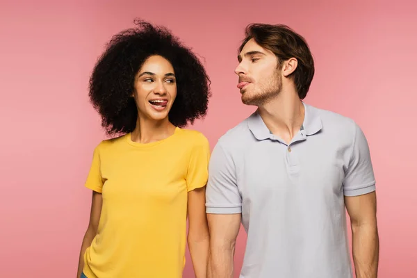 Alegre casal multiétnico olhando um para o outro e salientando línguas isoladas no rosa — Fotografia de Stock