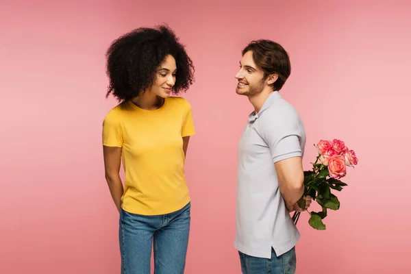 Curiosa mujer hispana cerca de hombre sosteniendo ramo de rosas detrás de la espalda aislado en rosa - foto de stock