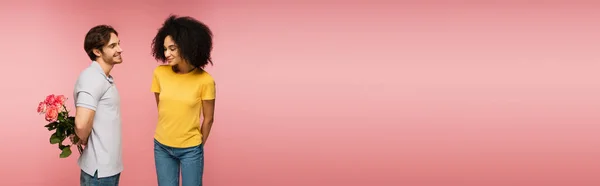 Young man holding roses behind back near smiling and shy hispanic woman isolated on pink, banner — Stock Photo