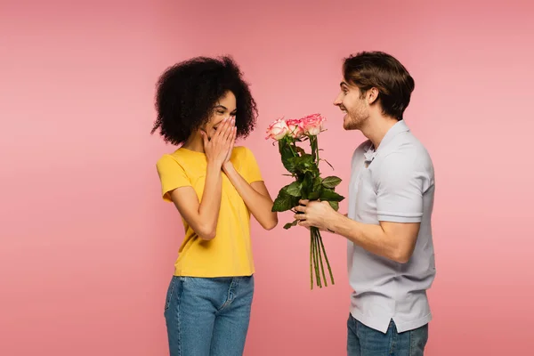 Веселий чоловік, що представляє квіти здивованій латинській жінці, що закриває обличчя руками ізольовано на рожевому — стокове фото