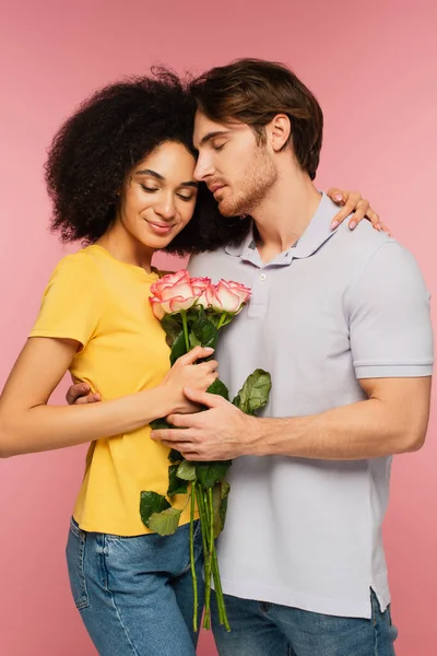 Feliz pareja interracial con ramo de rosas abrazando con los ojos cerrados aislados en rosa - foto de stock