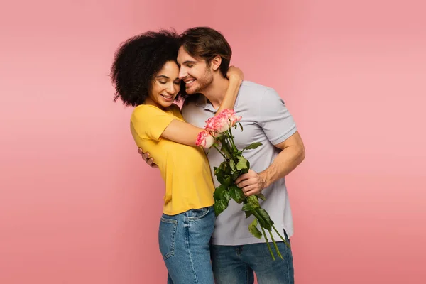 Lächelnder Mann mit einem Strauß Rosen, während er fröhliche hispanische Frau isoliert auf rosa umarmt — Stockfoto