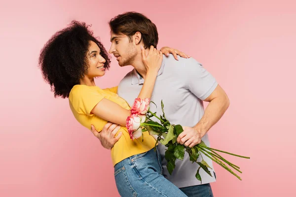 Junger Mann mit Blumen umarmt lächelnde lateinamerikanische Freundin isoliert auf rosa — Stockfoto