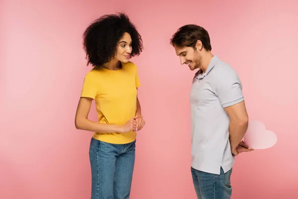 Mujer latina positiva y curiosa cerca del hombre tímido sosteniendo el corazón de papel detrás de la espalda aislado en rosa - foto de stock