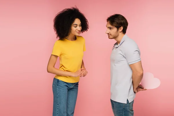 Jeune homme tenant coeur en papier derrière le dos près de curieuse petite amie hispanique isolé sur rose — Photo de stock