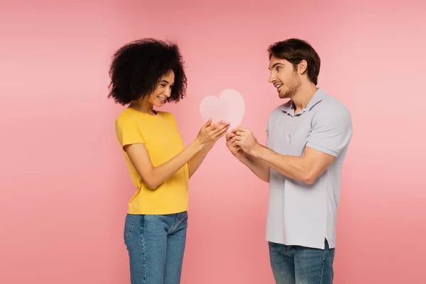 Homme présentant coeur de papier à jeune et jolie femme hispanique isolé sur rose — Photo de stock