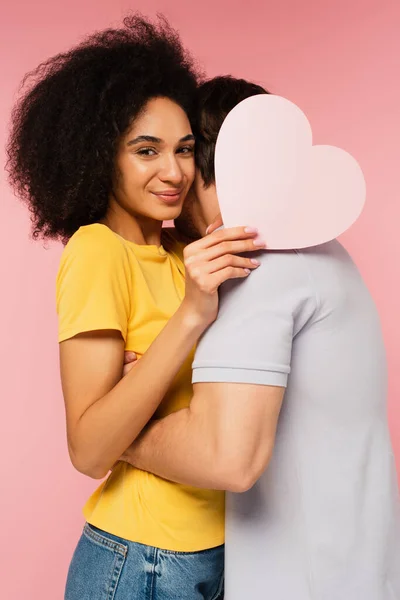 Mujer latina feliz con corazón de papel mirando a la cámara mientras su novio abrazándola aislado en rosa - foto de stock