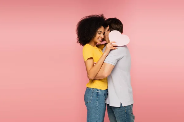 Jeune homme étreignant heureux femme latine tenant coeur en papier avec les yeux fermés isolé sur rose — Photo de stock