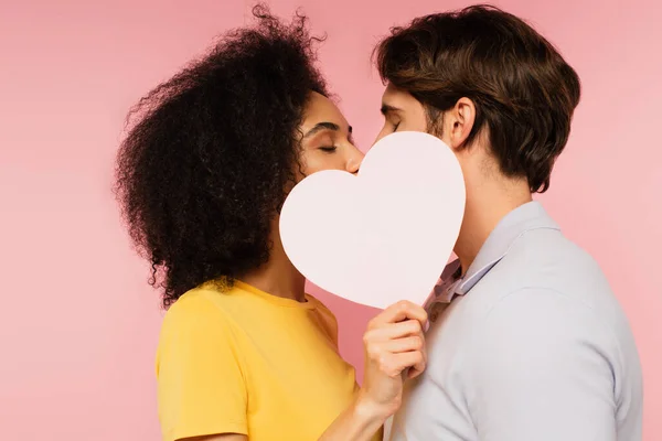 Casal interracial com olhos fechados beijando atrás de coração de papel em branco isolado em rosa — Fotografia de Stock