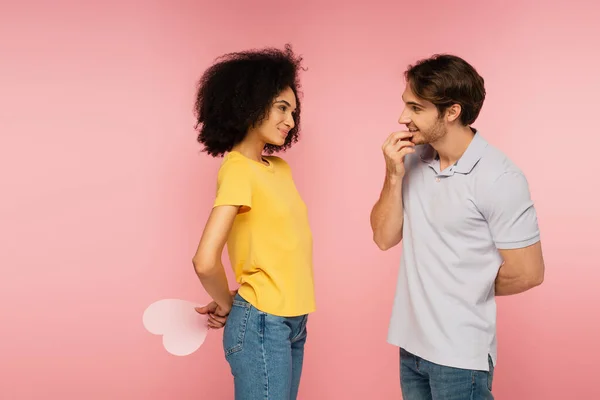 Gai hispanique femme tenant coeur en papier derrière le dos près petit ami curieux isolé sur rose — Photo de stock