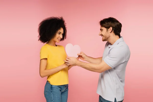 Uomo sorridente che presenta il cuore di carta alla ragazza latina soddisfatta isolato su rosa — Foto stock