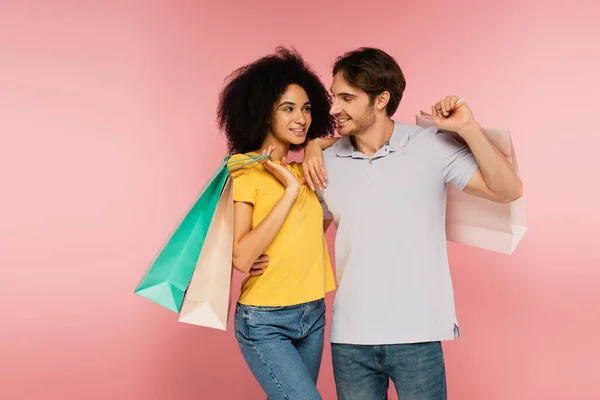 Feliz pareja interracial con bolsas de compras mirándose aislados en rosa - foto de stock