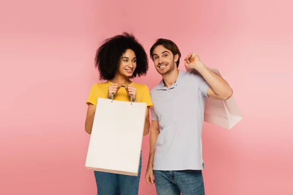 Alegre hombre sonriendo a la cámara cerca de la mujer hispana con bolsa aislada en rosa - foto de stock