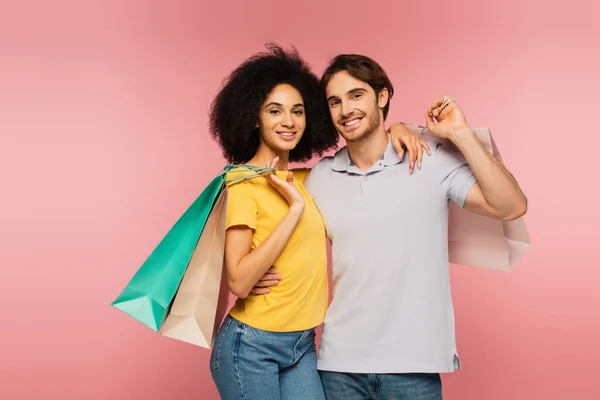 Pleased multiethnic couple with shopping bags hugging and smiling at camera isolated on pink — Stock Photo