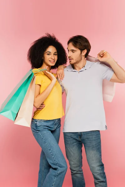 Joven con bolsas de compras mirando a la feliz novia hispana sosteniendo compras aisladas en rosa - foto de stock