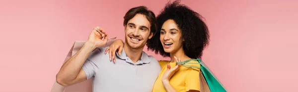 Glückliches multiethnisches Paar mit Einkaufstüten, die sich umarmen und isoliert auf rosa Transparenten wegschauen — Stockfoto