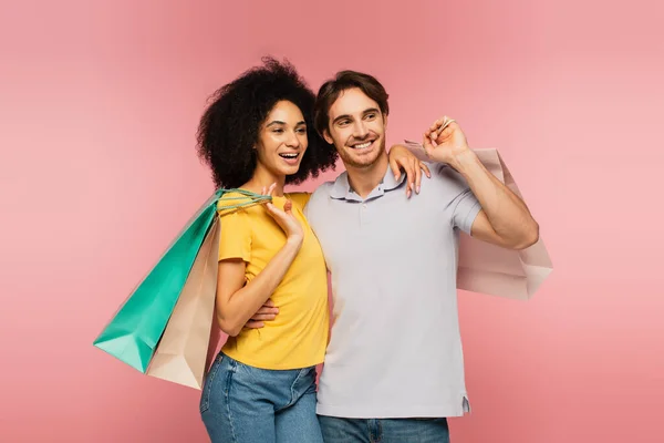 Joyeux couple interracial embrasser tout en tenant des sacs à provisions et regarder loin isolé sur rose — Photo de stock