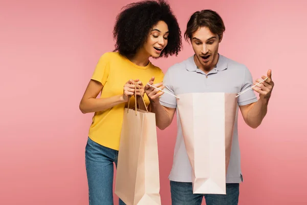 Surprised man looking into shopping bag near latin girlfriend isolated on pink — Stock Photo