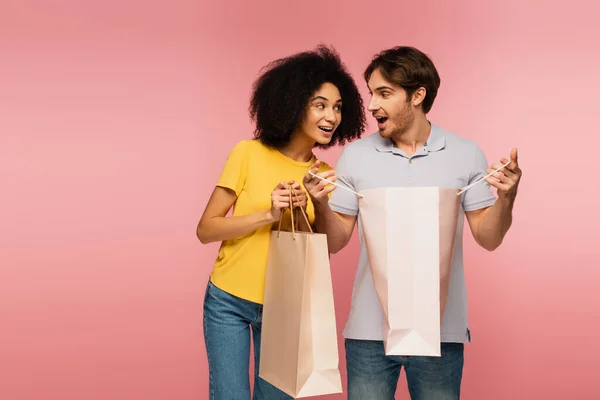 Asombrada pareja multiétnica con bolsas de compras mirándose aisladas en rosa - foto de stock