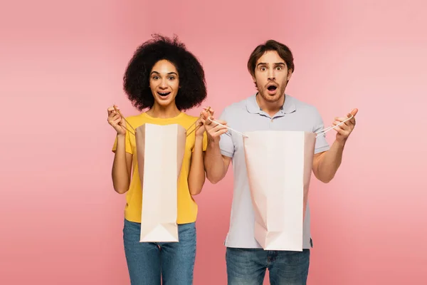 Verblüfftes multiethnisches Paar mit Einkaufstüten, während es isoliert in die Kamera auf rosa blickt — Stockfoto