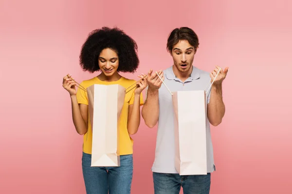 Felice donna ispanica in cerca di shopping bag vicino fidanzato stupito isolato su rosa — Stock Photo