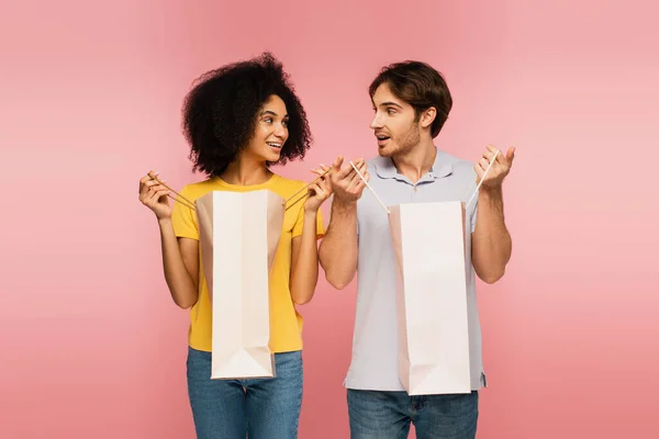Fröhliches gemischtrassiges Paar schaut einander beim Öffnen von Einkaufstüten isoliert auf rosa an — Stockfoto