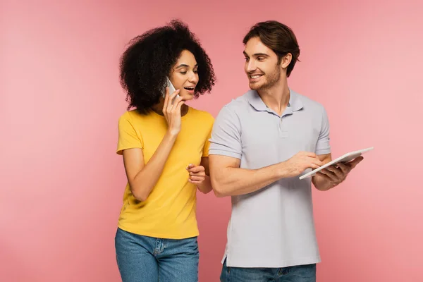 Allegra donna latina che parla su smartphone vicino sorridente uomo con tablet digitale isolato su rosa — Foto stock