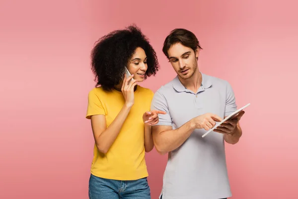 Mujer hispana hablando en smartphone y apuntando a la tableta digital junto con su novio aislado en rosa - foto de stock