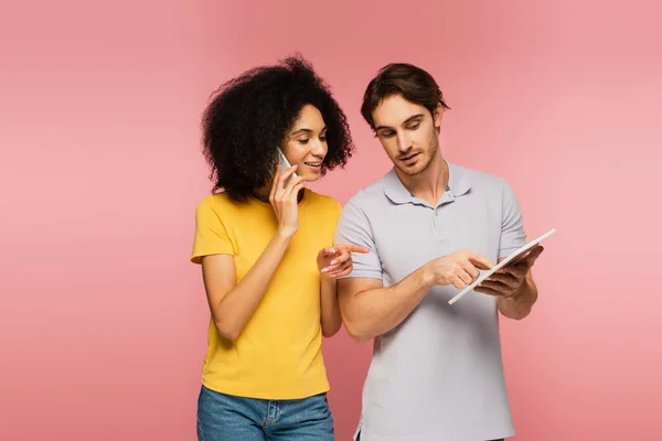 Spanisch Frau spricht auf Smartphone und zeigt auf digitales Tablet zusammen mit Freund isoliert auf rosa — Stockfoto