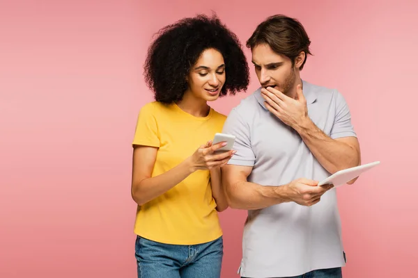 Smiling hispanic woman showing smartphone to shocked man with digital tablet isolated on pink — Stock Photo