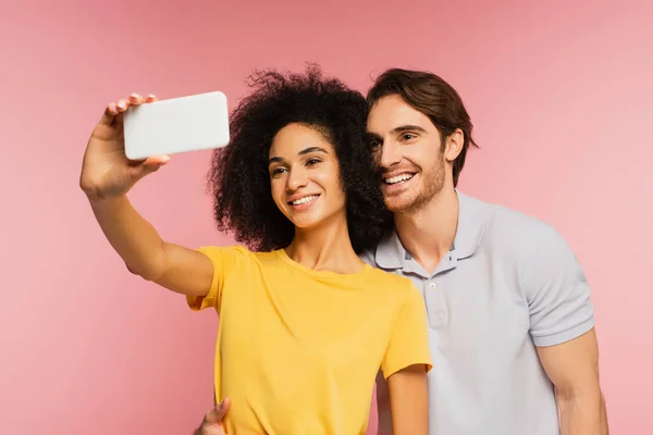 Joven sonriendo cerca alegre novia hispana tomando selfie en smartphone aislado en rosa - foto de stock