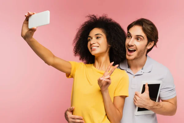 Homme étonné avec tablette numérique étreignant petite amie hispanique montrant signe de victoire tout en prenant selfie isolé sur rose — Photo de stock