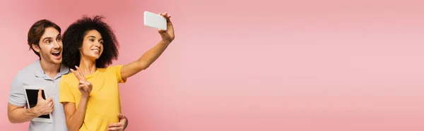 Счастливая латинская женщина показывает победный жест и делает селфи с изумленным мужчиной, держащим цифровой планшет изолирован на розовый, баннер — стоковое фото