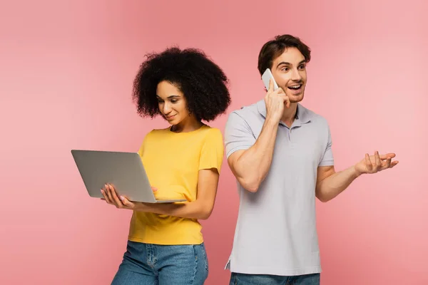 Fröhlicher Mann gestikuliert, während er auf dem Smartphone neben hispanischen Frau mit Laptop auf rosa isoliert spricht — Stockfoto