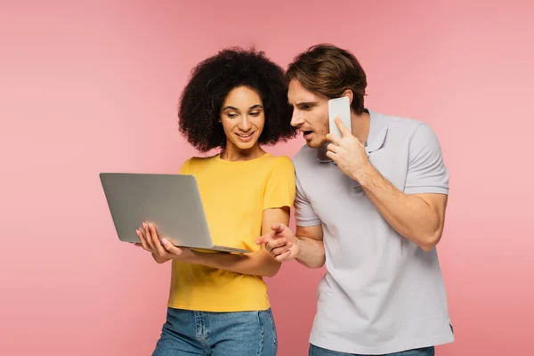 Homme surpris parlant sur téléphone portable et pointant vers l'ordinateur dans les mains d'une femme hispanique joyeuse isolée sur rose — Photo de stock