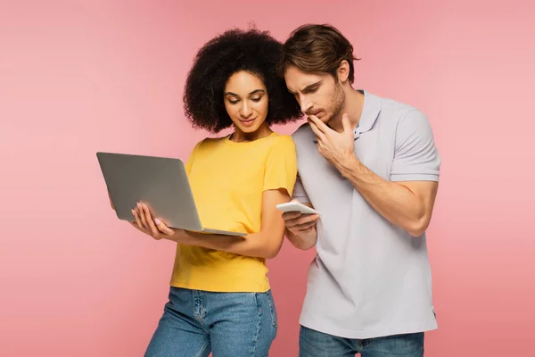 Nachdenklicher Mann schaut auf Handy neben hispanischer Frau mit Laptop isoliert auf rosa — Stockfoto